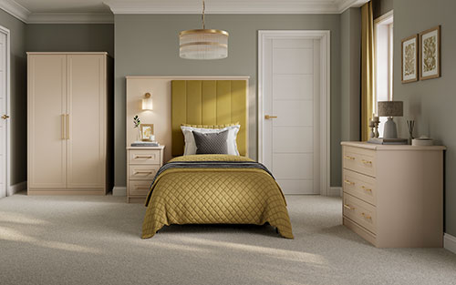 Beautifully designed care home bedroom featuring the Lincoln furniture collection in cashmere with champagne gold handles. The room includes a single bed with a mustard upholstered headboard, a matching nightstand, a spacious wardrobe, and a dresser. The décor is complemented by mustard and grey accents, creating a warm and inviting atmosphere.