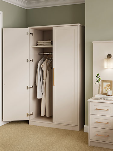 Lincoln wardrobe with the left-hand door open showing garments hanging in the wardrobe, and shirts folded on the top shelf. Right hand door is closed. Next to it is the corner of the chest of drawers.