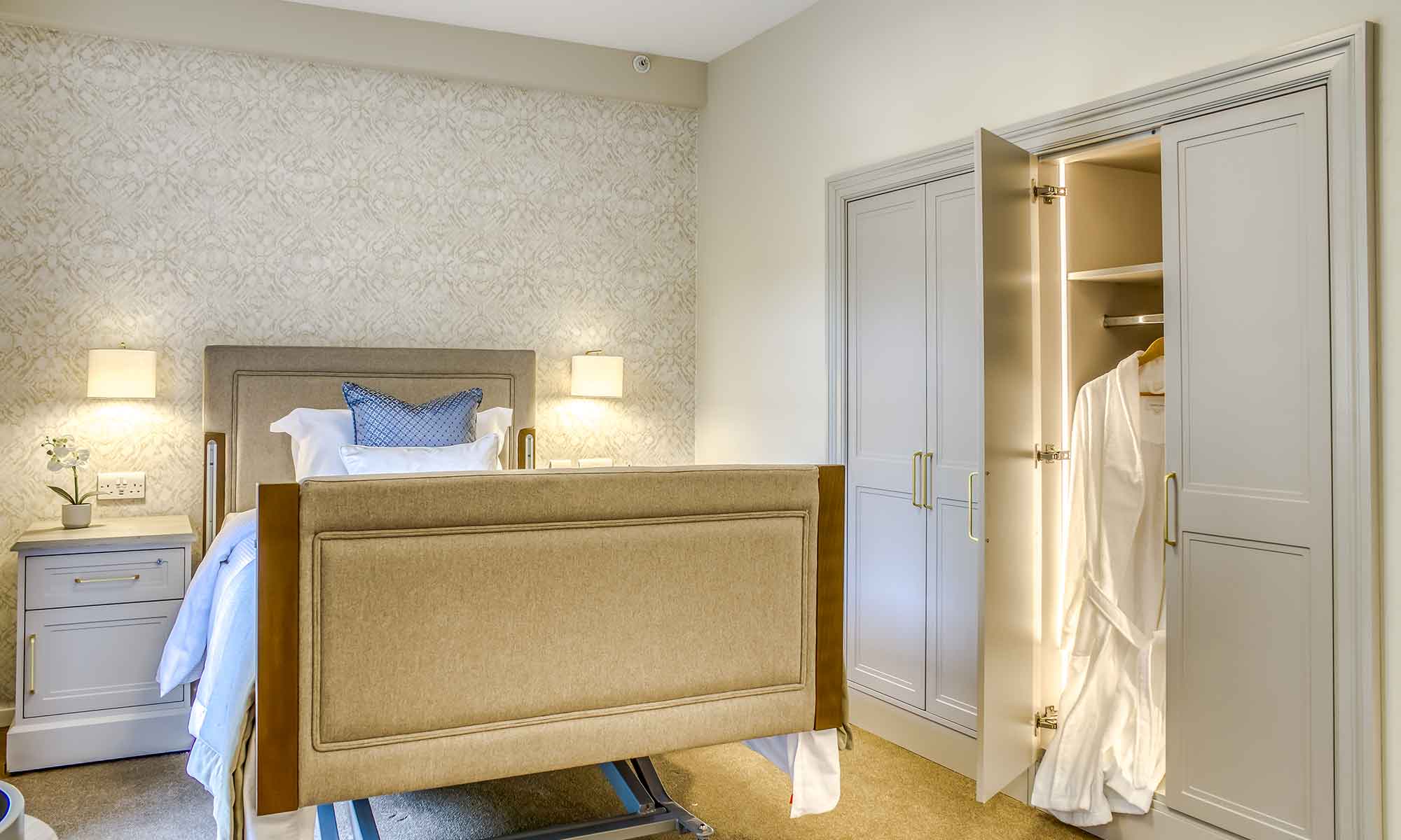 Luxurious bedroom featuring an upholstered adjustable bed with a soft headboard, flanked by elegant bedside cabinet and built-in fitted wardrobes, offering a comfortable and stylish living space designed for senior care.