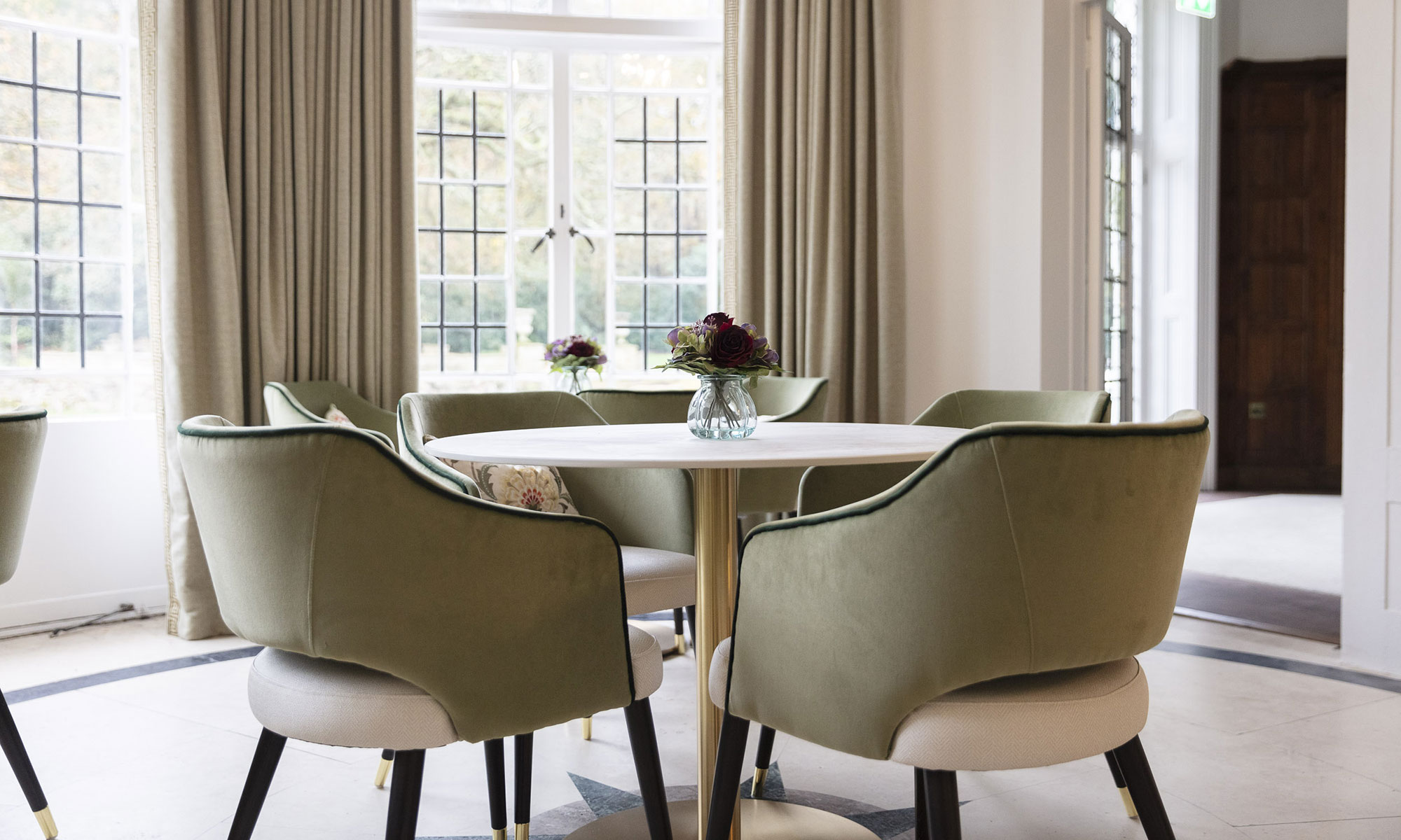 Upper Court Care Home - dining room. A stylish dining area featuring a round table with plush green velvet chairs, elegant gold accents, and a floral centerpiece. Large windows with classic leaded glass panels and soft neutral curtains allow natural light to enhance the sophisticated setting.